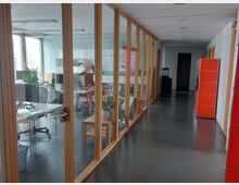 Ein Büroflur mit gläsernen Trennwänden und Holzelementen, die in einen hellen Besprechungs- und Arbeitsbereich mit Schreibtischen und Pflanzen führen. Auf der rechten Seite befinden sich rote Aktenschränke, der Boden ist dunkel und glänzend.