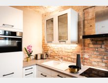 Moderne Küche mit Backsteinwand, weißen Schränken und grauer Arbeitsplatte. Sichtbare Elemente sind ein Einbauofen, eine Spüle, Glasfront-Hängeschränke, eine Dunstabzugshaube, eine Orchideenpflanze sowie Küchenutensilien wie Wasserkocher und Schüsseln.