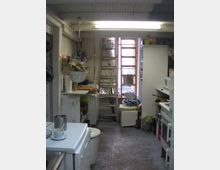 Eine kleine, schmale Küche mit offenen Regalen, einer Arbeitsfläche und einem Leiterregal im Hintergrund. Die Decke ist mit weißen Holzpaneelen verkleidet, und es gibt eine helle Fensterfront am Ende des Raums, die etwas natürliches Licht hereinlässt.