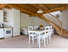 Gemütlicher Wohnraum mit rustikalen Holzbalken und Terrakottafliesenboden, ausgestattet mit einer offenen Küchenzeile, Esstisch mit weißen Stühlen und einer geschwungenen Holztreppe, die zu einem oberen Bereich führt. Im Hintergrund befindet sich ein weißes Sofa vor einer hellen Wand.