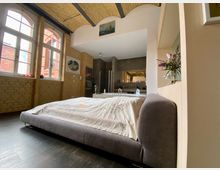 Geräumiges Schlafzimmer in einem Altbau mit sichtbaren Ziegelwänden und großen Rundbogenfenstern. Im Vordergrund steht ein graues Polsterbett, im Hintergrund ist ein separates Badbereich mit Waschbecken und Spiegel sichtbar.