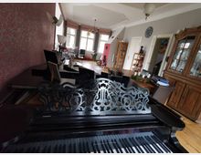 Ein Wohnraum mit einem verzierten schwarzen Flügel im Vordergrund, einem Holzesstisch und Stühlen in der Mitte sowie einem großen hölzernen Vitrinenschrank an der rechten Wand. Im Hintergrund sind große Fenster, ein Kronleuchter und eine teppichbedeckte Sitzecke sichtbar.