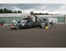 Ein Parkplatz vor einer Industriehalle mit Glasfassaden und den Beschriftungen "C1" und "C2". Im Vordergrund ein Filmset mit einem grauen Transporter, einem Kamerakran, mehreren Personen und technischem Equipment.