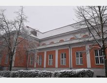 Ein zweistöckiges Gebäude mit orange-weißer Fassade und großen rechteckigen Fenstern, umgeben von schneebedeckten Bäumen und einer Hecke im Vordergrund. Der Himmel ist grau, und es schneit.