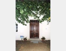 Eine braune Holztür mit kunstvollen Verzierungen befindet sich in einer hellen, verputzten Fassade. Vor der Tür stehen zwei Blumentöpfe, während herabhängende Äste mit grünen Blättern den oberen Bereich des Bildes einrahmen.