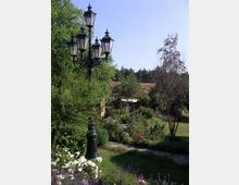 Ein Garten mit Blumenbeeten, Sträuchern und einem gepflasterten Weg. Im Vordergrund steht eine schwarze, dekorative Laterne mit mehreren Lampen, im Hintergrund ist ein ziegelgedecktes Gebäude teilweise sichtbar, umgeben von Bäumen.