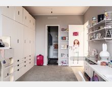 Kinderzimmer mit einer grauen Wand und einem Hochbett links. Ein großer Einbauschrank nimmt die linke Wand ein, während an der rechten Wand ein Schreibtisch und offene Regale mit Spielzeug und Büchern stehen. Die Tür in der Mitte des Raumes ist mit einer Kinderzeichnung dekoriert.