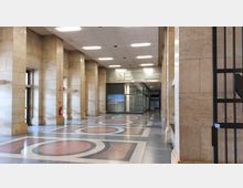 Großer, heller Innenraum mit hohen hellen Steinwänden und rechteckigen Säulen. Der Boden ist mit geometrischen Mustern in rot, schwarz und grau gestaltet, in der Mitte hängt eine analoge Wanduhr, und im Hintergrund befinden sich Glastüren und ein Notausgangsschild.