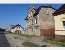 Eine Straße mit mehrheitlich kleinen Wohnhäusern, darunter ein verfallenes, mehrstöckiges Gebäude mit bröckelnder Fassade und dekorativen Stuckelementen. Im Vordergrund sind ein gepflasterter Gehweg, ein schmiedeeisernes Geländer und eine Zufahrt mit unbefestigtem Untergrund zu sehen.