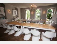 Ein heller und stilvoll eingerichteter Seminarraum mit langen, hellen Holztischen, weißen Kunststoffstühlen und einem Kronleuchter. Große Fenster mit Rundbögen bieten Blick auf grüne Bäume, während Pflanzen, Geschirr und Wasserflaschen die Tische dekorieren.