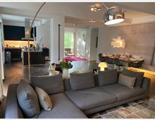 Geräumiges Wohnzimmer mit moderner Einrichtung und offener Küche im Hintergrund. Dunkler Holzboden, graues Sofa, Esstisch mit gedeckten Stühlen und ein eleganter Lampenschirm schaffen ein stilvolles Ambiente.