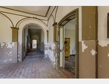 Ein verlassener Innenraum mit einem langen Flur, gekennzeichnet durch abblätternde Farbe an den Wänden und einen abgenutzten Fliesenboden. Der Raum rechts zeigt einen alten, gelbfarbenen Kachelofen, während am Ende des Flurs ein geöffneter Durchgang ins Freie sichtbar ist.