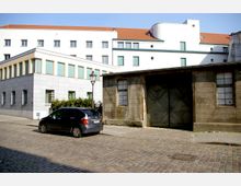 Straßenszene mit einem modernen weiß verputzten Gebäude mit Flachdach und großen Fenstern im Hintergrund sowie einem älteren, verwitterten Gebäude aus Stein mit Holztür und vergittertem Fenster im Vordergrund. Ein geparktes Auto steht auf dem gepflasterten Straßenabschnitt.