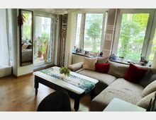 Helles Wohnzimmer mit Ecksofa, roten und gestreiften Kissen sowie einem weißen Couchtisch mit Tischdecke und Dekoration. Große Fenster bieten Blick auf einen Garten, und links führt eine Glastür zu einem Balkon mit Pflanzen.
