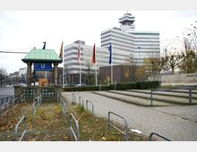 U-Bahn-Eingang Theodor-Heuss-Platz mit grünem Pavillondach und einem Schild über dem Eingang, flankiert von Fahnenmasten. Im Hintergrund ein mehrstöckiges, modernes Gebäude mit gestreifter Fassade, ein Plattenweg und kahle Bäume umrahmen die Szene.