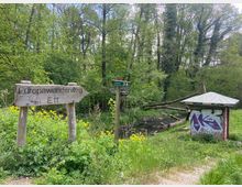 Holzwegweiser mit der Aufschrift „Europawanderweg E11“ in einer bewaldeten Umgebung mit dichtem Grün, einem kleinen Bach und einem mit Graffiti besprühten Unterstand mit Spitzdach. Im Hintergrund sind Bäume und vereinzelte umgestürzte Stämme zu sehen.
