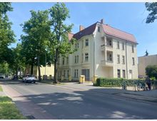 Wohnstraße mit einem hellen, dreigeschossigen Wohnhaus mit roten Dachziegeln und zwei kleinen Balkonen. Links sind Bäume entlang der Straße und geparkte Autos sichtbar, die Umgebung wirkt ruhig und grüner Wohngegend zugehörig.