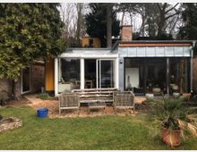 Flacher Wohnhausanbau mit gelb gestrichenen Wänden, großen Glasfenstern und einem Schornstein aus Backstein. Im Vordergrund befindet sich ein Garten mit Sitzbänken, Topfpflanzen und einem kleinen Rasenbereich, umgeben von Bäumen und Sträuchern.