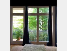 Großes Fenster in einem modernen Raum mit dunklen Vorhängen, das auf einen Balkon führt, der mit einer Pflanze dekoriert ist, und im Hintergrund grüne Bäume zeigt. Der Boden ist aus Holz, und im Vordergrund ist ein Bett mit einer hellen Decke sichtbar.