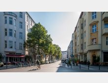 Straßenszene in einem Wohnviertel mit mehrstöckigen Gebäuden im Altbaustil auf beiden Seiten, links ein Café mit roten Markisen und Sitzplätzen im Freien, rechts ein gelb gestrichenes Gebäude mit verzierten Fassadenelementen. Bäume säumen die Straße, Fahrräder und Fußgänger sind zu sehen, und parkende Autos stehen entlang des Bürgersteigs.