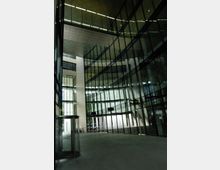 Großes, modernes Atrium mit hohen Glasfassaden, die die umliegende Architektur spiegeln. Im Vordergrund befindet sich ein runder gläserner Dreh-Eingang, während im Hintergrund Fahrräder an einer Wand lehnen.
