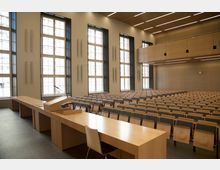 Ein moderner Hörsaal mit mehreren Reihen aus Holz- und Kunststoffsitzen, großen Fenstern mit Sprosseneinteilung und klaren Linien. Vorne befindet sich ein Dozententisch mit Rednerpult, Mikrofon und Stuhl; die Wände und Decke sind mit Holzpaneelen verkleidet.