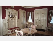 Ein klassisch eingerichteter Raum mit roten Wänden, cremefarbenen Türen und holzverzierten Elementen. In der Mitte steht ein runder Marmortisch mit einem Kerzenhalter, umgeben von gepolsterten Stühlen mit rot-weiß gestreiftem Stoff, ergänzt durch Vorhänge in ähnlichem Design.