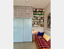 Ein kleines Zimmer mit einem Tagesbett, das mit einer karierten Decke und bunten Kissen dekoriert ist. Links steht ein großer, hellblauer Schrank mit Schiebetüren, rechts daneben befindet sich ein Regal mit Büchern an der Wand und eine Tischlampe auf einer darunterliegenden Fläche.