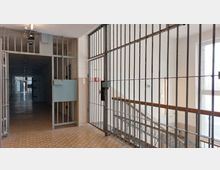 Ein Flur in einem Gefängnis mit vergitterten Türen und Fenstern, einer Metalltreppe auf der rechten Seite und einem Korridor, der in der Tiefe weitere Zellentüren zeigt. Der Boden ist gefliest, und die Wände wirken schlicht und funktional gestaltet.