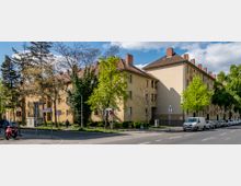 Mehrfamilienhäuser mit hellgelber Fassade und roten Ziegeldächern an einer Straßenecke, umgeben von Bäumen und geparkten Autos entlang des Gehwegs. Die Szenerie befindet sich bei Tageslicht und zeigt eine ruhige Wohngegend.