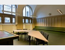 Ein heller Raum mit gewölbter Decke, großen Fenstern mit vergittertem Glas, einer holzverkleideten Wand und einer grünen Sitzbank. Im Zentrum stehen mehrere zusammengestellte, geometrische Tische und gepolsterte Stühle.