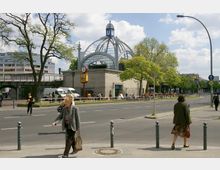 Straßenszene mit einem U-Bahn-Eingang in einem historischen Gebäude mit einer markanten Kuppel aus Glas und Metall. Im Vordergrund Gehweg und Straße, im Hintergrund Bäume, Passanten und Sitzmöglichkeiten vor dem Gebäude.