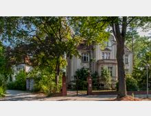 Villenartiges Gebäude namens "Haus Holstein" mit dekorativer Fassade, großen Fenstern und rotem Ziegeldach, umgeben von dichtem Grün und hohen Bäumen. Im Vordergrund ein schmiedeeisernes Tor mit Backsteinpfosten, das zu einer ruhigen Straße führt.