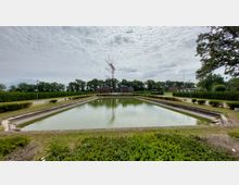 Großes rechteckiges Wasserbecken, umgeben von gepflegten Grünflächen und Hecken, im Hintergrund Bäume, mehrere Backsteingebäude und eine hohe Funkantenne. Der Himmel ist bewölkt, und die Szene liegt in einer offenen, parkähnlichen Umgebung.
