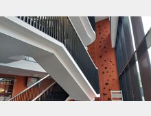 Innenaufnahme eines modernen Gebäudes mit einer frei hängenden Treppe und schwarzen Geländern. Im Hintergrund ein orangefarbener Kletterbereich mit Griffen und Kletterseilen, daneben eine große Fensterfront.