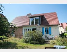 Ein Einfamilienhaus mit rotem Satteldach und teils weiß verputzter, teils roter Backsteinfassade. Im Vordergrund befindet sich ein Garten mit Sträuchern, Wiese und einem kleinen Sitzbereich mit Tisch und Stühlen.