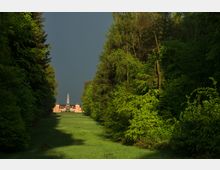 Eine Allee mit dicht bewachsenen, grünen Bäumen führt zu einem historischen Gebäude mit einer zentralen Turmspitze. Im Vordergrund liegt eine Wiese, während der Himmel dunkel bewölkt ist und das Sonnenlicht die Szenerie akzentuiert.