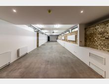 Langer, schlichter Raum mit Betonboden, weiß gestrichenen Wänden und freiliegenden Klinkersteinen an einer Seite. Die Decke zeigt sichtbare Lüftungsrohre und moderne Beleuchtung, an den Wänden sind Heizkörper und Lüftungsgitter angebracht.