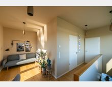 Helles Wohnzimmer oder Flur mit minimalistischer Einrichtung, bestehend aus einer grauen Couch, mehreren Pflanzen, Holzfußboden und weißen Türen. Wände sind beige gestrichen und mit dekorativen Bildern und moderner Beleuchtung ausgestattet.