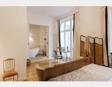 Helles Schlafzimmer mit Holzboden, einem Bett mit Holzrahmen und einer großen Fensterfront mit cremefarbenen Vorhängen. Im Hintergrund ein offenes Badezimmer mit freistehender Badewanne, daneben ein kleiner Schreibtisch und ein Raumtrenner.