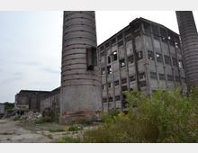Abrissreifes Industriegebäude mit zerbrochenen Fenstern und stark gealterter Fassade, flankiert von zwei hohen Ziegelschornsteinen. Im Vordergrund sind Gestrüpp und Bauschutt zu sehen, die den verwahrlosten Zustand der Umgebung unterstreichen.