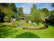 Ein Park mit einer kleinen Wasserbahn, auf der grüne Boote mit Menschen fahren. Im Hintergrund sind Schilf, ein weißes Büsten-Denkmal, ein Blockhaus und Bäume zu sehen.