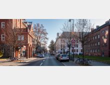 Straßenszene in einem städtischen Bereich mit mehrstöckigen Ziegelgebäuden auf beiden Seiten. Die Straße ist gesäumt von unbelaubten Bäumen, parkenden Autos und Fahrrädern; im Hintergrund steht ein weißes Gebäude mit einem kleinen Turm.