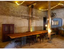 Ein rustikaler Innenraum mit unverputzten Ziegelwänden, einem langen Holztisch mit Holzstühlen und einer Holzbalkendecke. An der Wand hängen Dekorationen wie ein Fahrrad, und eine Lichterkette schmückt die Decke.