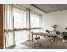 Heller Raum mit großer Fensterfront und transparenten Vorhängen, bestehend aus einer Massageliege, einem kleinen Tisch mit zwei Stühlen sowie Pflanzen in weißen Töpfen. Der Boden ist aus Beton, teils mit einem beige-farbenen Teppich, und die Decke besteht aus sichtbarem Beton.