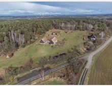 Luftaufnahme eines Freilichtmuseums mit mehreren historischen Fachwerkhäusern und Reetdachgebäuden, umgeben von Wiesen und einem angrenzenden Wald. Im Hintergrund sind Windräder und Felder zu sehen, während eine Landstraße durch die Landschaft führt.