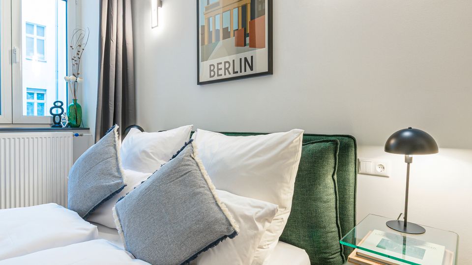 Modernes Schlafzimmer mit grün gepolstertem Bett, weißer Bettwäsche und mehreren Kissen, daneben ein gläserner Nachttisch mit schwarzer Lampe. Helle Wände mit zwei Wandleuchten und einem „Berlin“-Poster, großes Fenster mit grauem Vorhang sowie Holzboden.