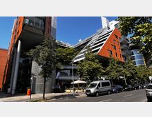 Moderne Gebäudearchitektur mit einer schrägen Fassade aus orangefarbenem Material und großen Glasflächen, umgeben von Bäumen. Im Vordergrund verläuft eine Straße mit geparkten Autos und einem Bürgersteig.