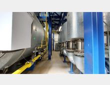 Industrieanlage mit einem großen Metalltank auf der linken Seite, verbunden mit gelben Rohrleitungen, sowie mehreren silbernen Rohren und Verbindungsstücken auf der rechten Seite. Die Struktur ist von blauen Stahlträgern und einer Gitterplattform im oberen Bereich umgeben.