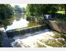 Ein kleiner Fluss mit einer niedrigen Staustufe, über die Wasser fließt, umgeben von üppigen Bäumen und einem betonierten Ufer; im Hintergrund ist eine ruhige Flusslandschaft und ein parkähnliches Gebiet zu sehen.